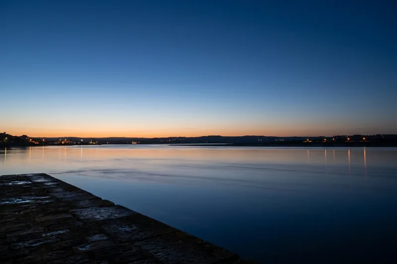 Blue hour over the Tagus estuary, the far shore lit in sodium orange under a cobalt sky.
